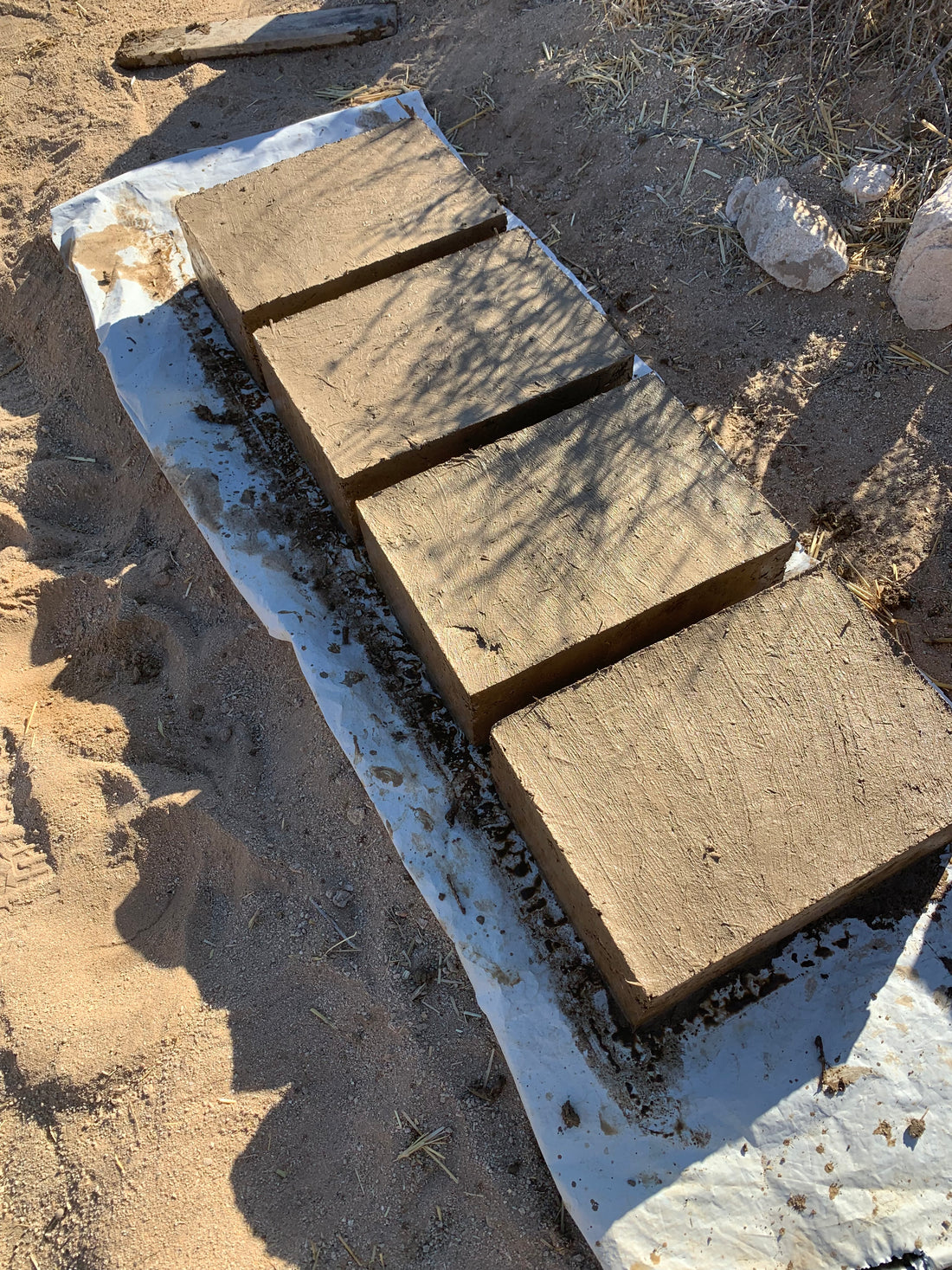 The image shows four perfectly square adobe blocks lined up in a row after being taken out of the mold. Made from clay, hay, and straw and are shown on a plastic sheet improving the drying process and protecing them from the sand.