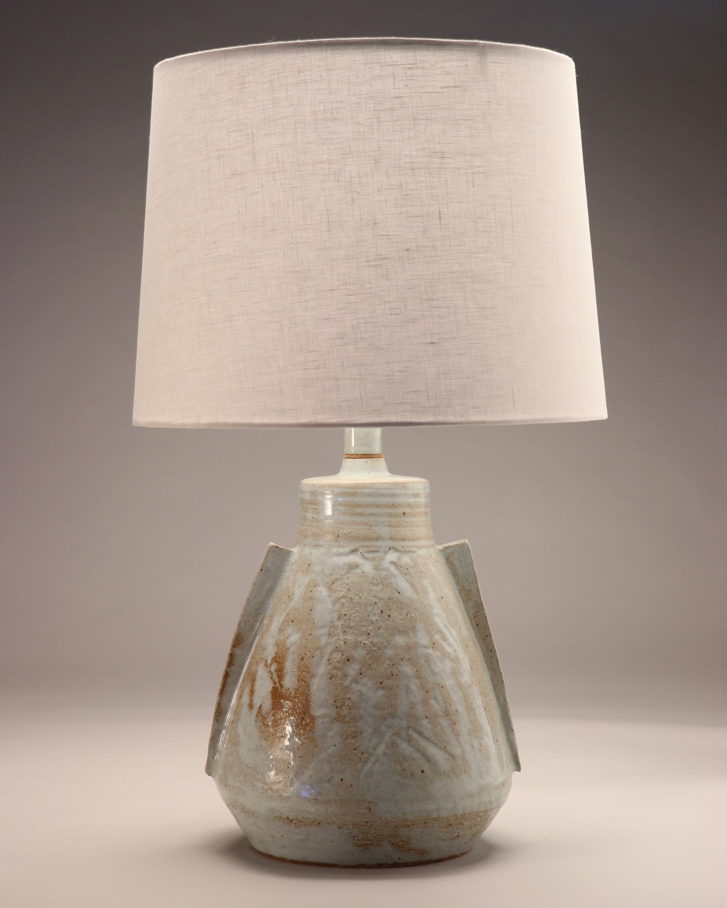 Image shows a modern abstract lamp made from ceramic with a white linen lamp shade. The lamp base is a grey-blue color with gold detailing. To the rounded but angular body two angular handles are attached.