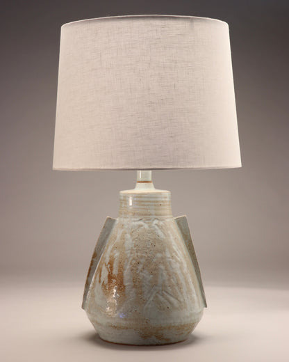 Image shows a modern abstract lamp made from ceramic with a white linen lamp shade. The lamp base is a grey-blue color with gold detailing. To the rounded but angular body two angular handles are attached.