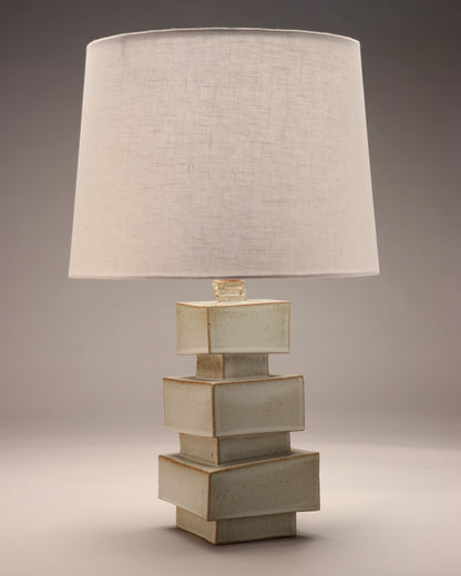 The image shows an abstract geometric lamp base with a white linen shade. Three large blocks are stacked with small blocks separating the layers. The body is a grey-blue color with brown or gold glaze showing through the edges. 