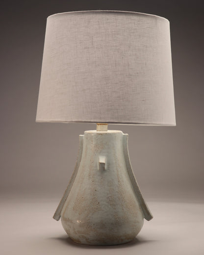 The image shows a modernistic lamp base with a white linen lamp shade.  The shape has elements reminiscent of an abstract bird. The colors are a pattern of tan and very light blue.