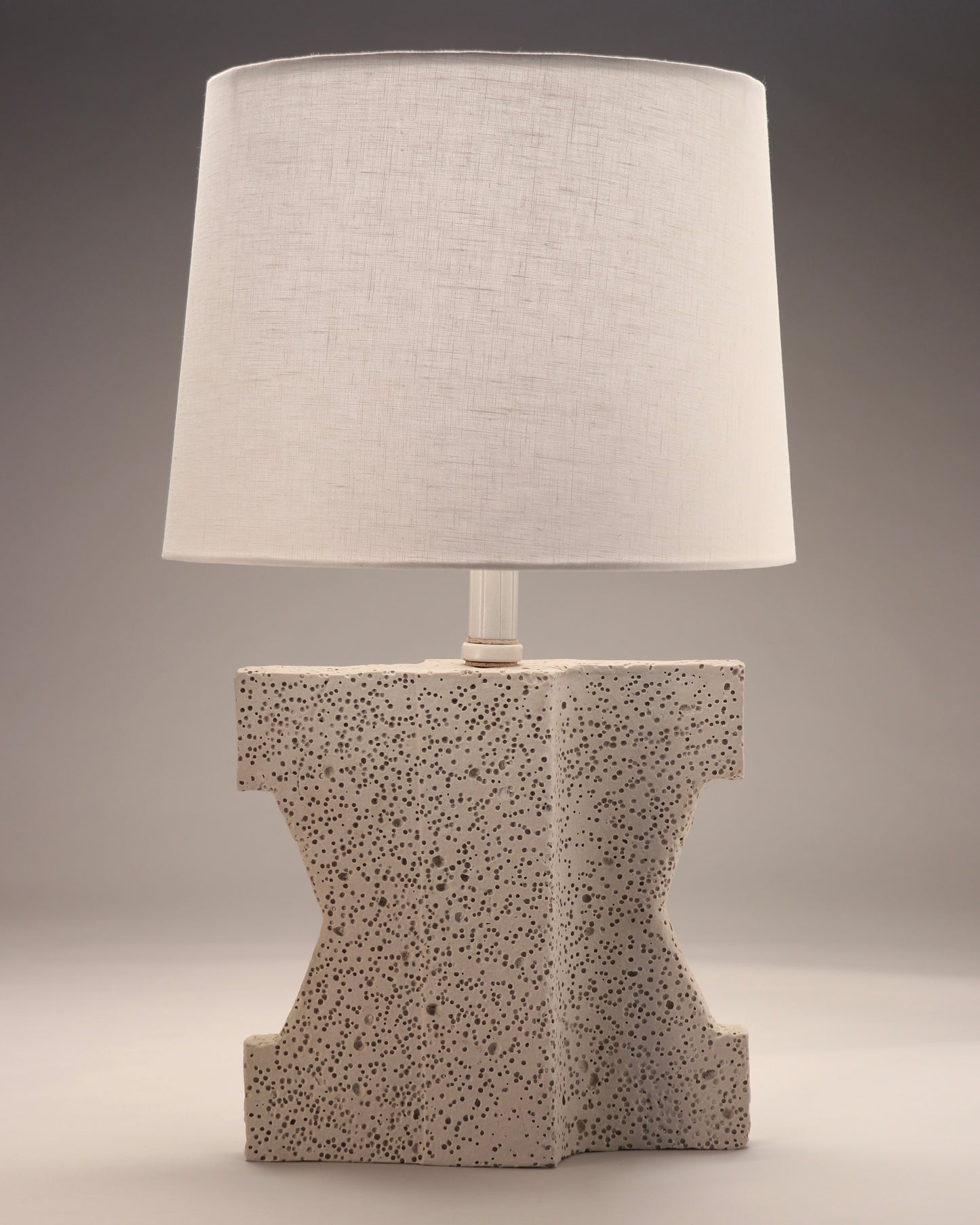 The image shows an abstract lamp base with a white linen shade. There is a very unique angled brick shape with a porous texture. The lamp is a creamy white.