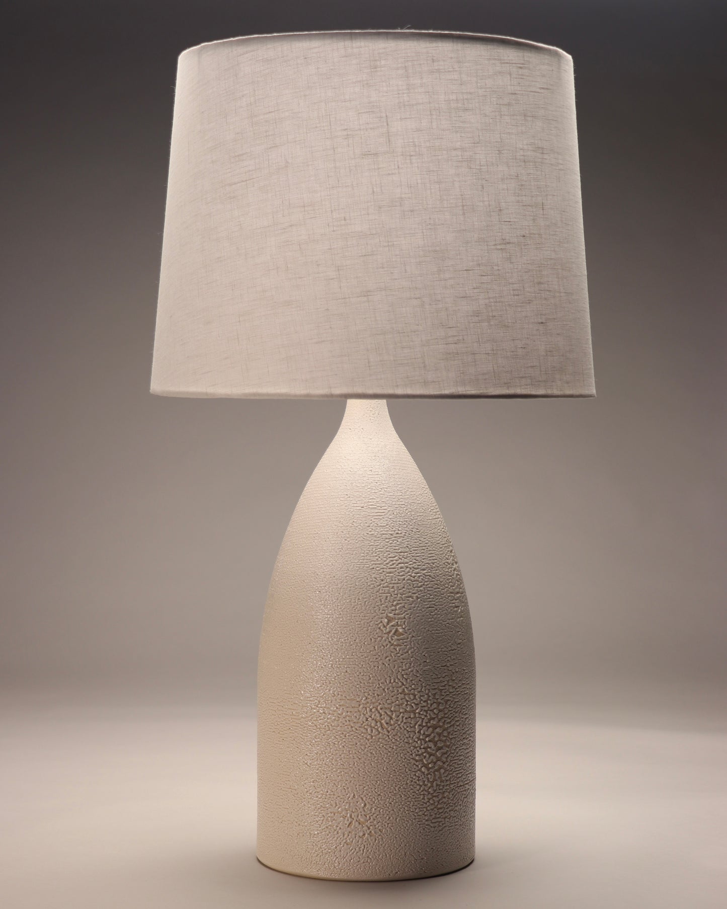 The image shows a modern and minimalist style lamp with a white linen shade. It is a tall and oval cylinder. There is a very unique and detailed texture on the piece made rom glaze and printed layers.