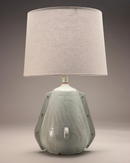 The image shows a light blue lamp shade with grey accents and a white linen shade. The style is Rustic and abstract with modern elements. Two handle-like ribs are smoothed into the side with holes punctured for an added element. At the front of the piece is a small circular ridge where another hole is punctured through.