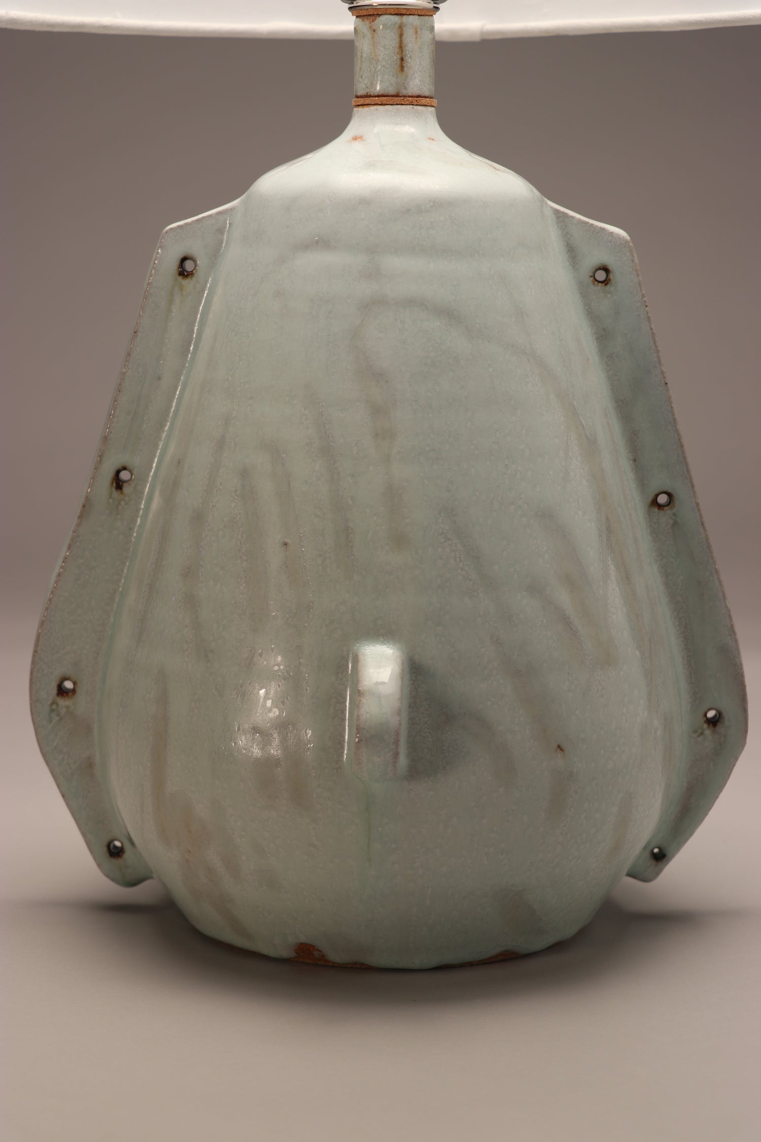 The image shows a light blue lamp shade with grey accents. The style is rustic and abstract with modern elements. Two handle-like ribs are smoothed into the side with holes punctured for an added element. At the front of the piece is a small circular ridge where another hole is punctured through.