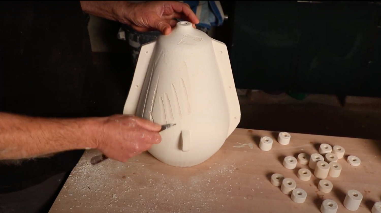 Screen shot depicting the process of making the lamp "Queenstown." The image shows the lamp base after the first glaze where an abstract design is being carved into the front. The glaze looks like a powdery white before it is fired into a grey and blue color.