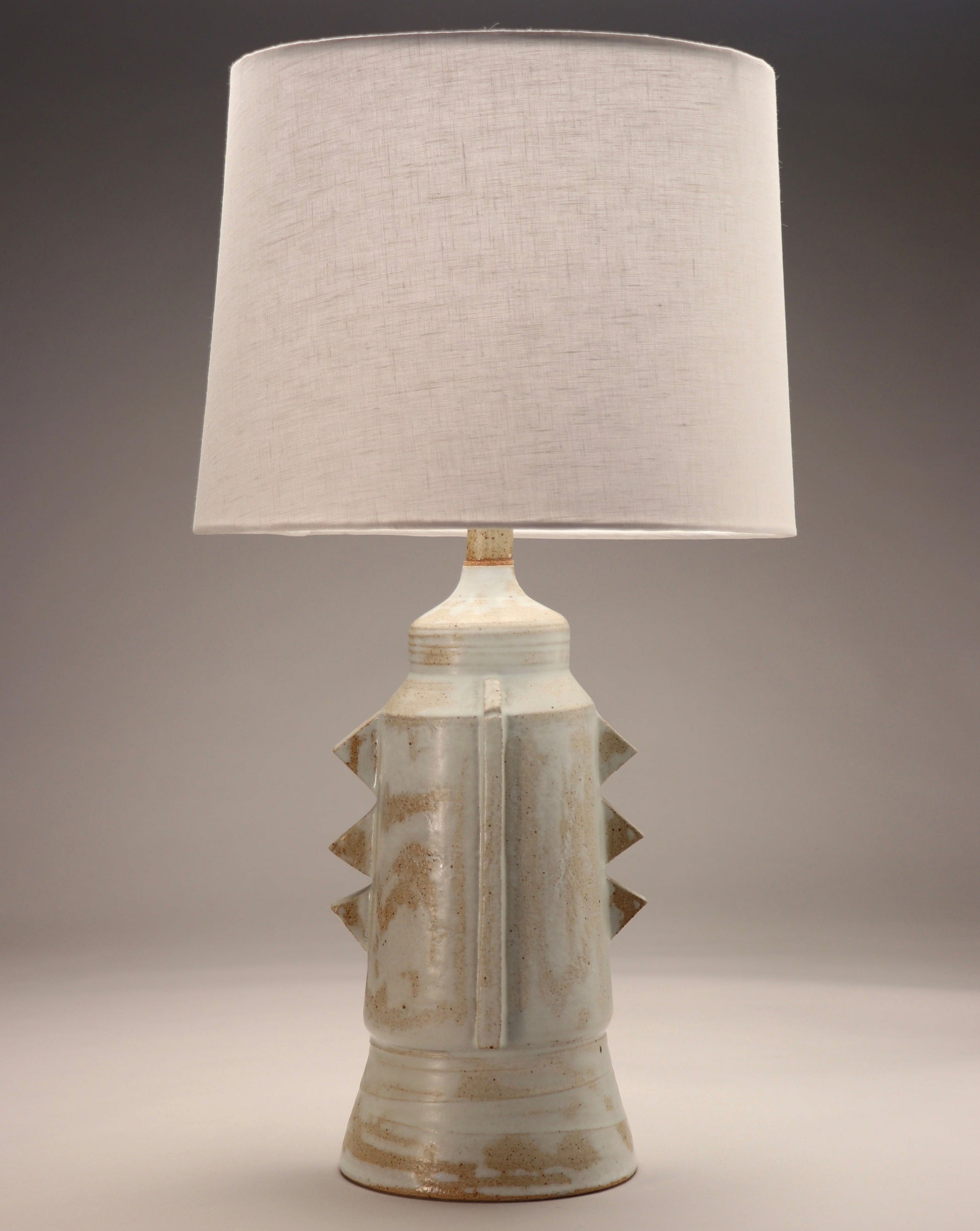 Image shows a cylindrical lamp base accompanied by a white linen shade. Rustic angular accents are added t the body of the piece. Earthy toned glazes were used for this piece.